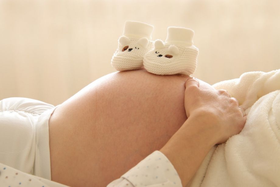 Gesellschaftliche Erwartungen - liegende schwangere Frau, die auf dem Rücken liegt und ihren Bauch zeigt. Obenauf stehen zwei gehäkelte Babyschuhe.