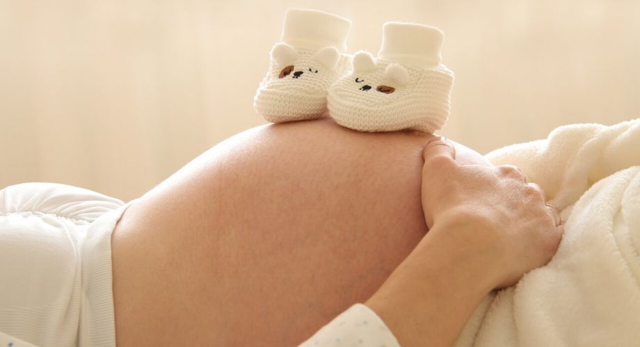 Gesellschaftliche Erwartungen - liegende schwangere Frau, die auf dem Rücken liegt und ihren Bauch zeigt. Obenauf stehen zwei gehäkelte Babyschuhe.